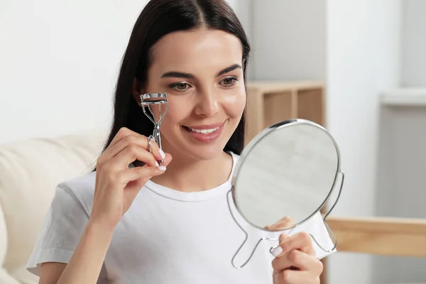 Beautiful young woman with mirror using eyelash curler indoors
