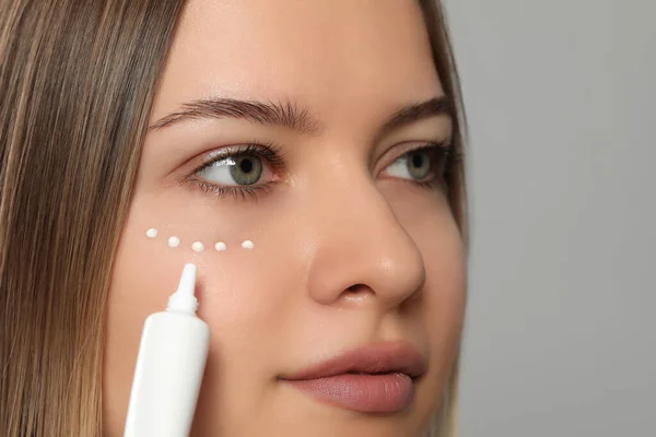 Mujer Joven Aplicando Crema Bajo Los Ojos Sobre Fondo Gris —  Fotos de Stock