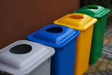 Açık havada kahverengi duvarın yanında birçok renk geri dönüşüm kutusu var.