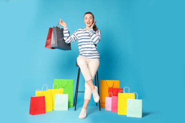 Heyecanlı bir kadın açık mavi arka planda renkli alışveriş torbaları taşıyor.