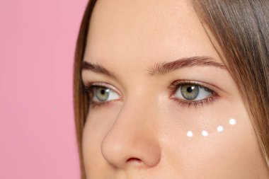 Young woman with cream around eye on pink background, closeup