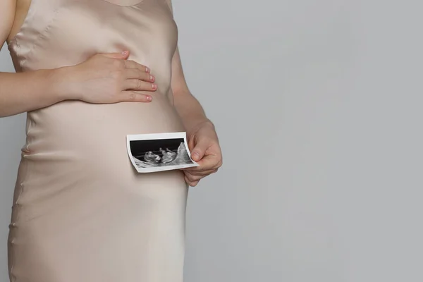 Pregnant woman with ultrasound picture of baby on light grey background, closeup. Space for text