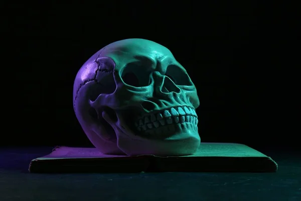 Human skull on book in colorful neon lights against black background