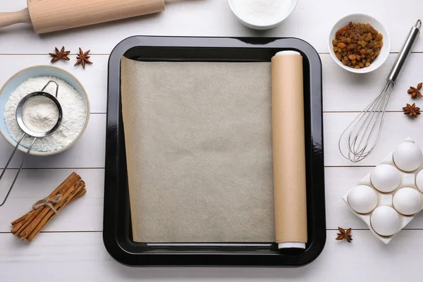 Baking pan with parchment paper, different ingredients and kitchen tools on white wooden table, flat lay