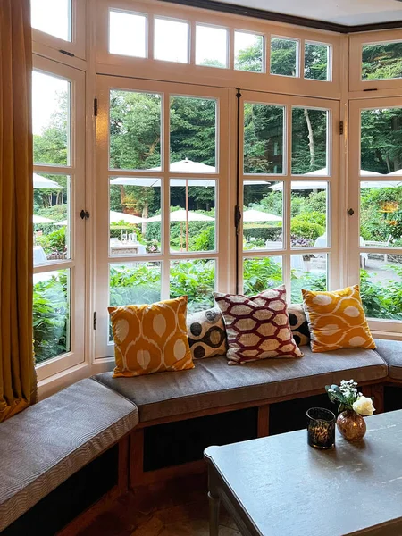Wooden table near bay window seat with cushions in cozy room. Interior design