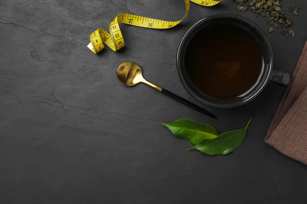 Cup of herbal diet tea and measuring tape on black table, flat lay with space for text. Weight loss concept