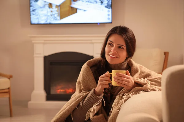 Young woman with cup of tea relaxing near fireplace at home