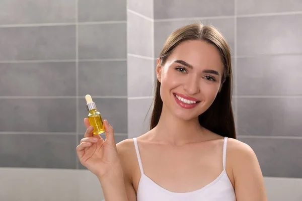 Young woman with bottle of essential oil in bathroom, space for text