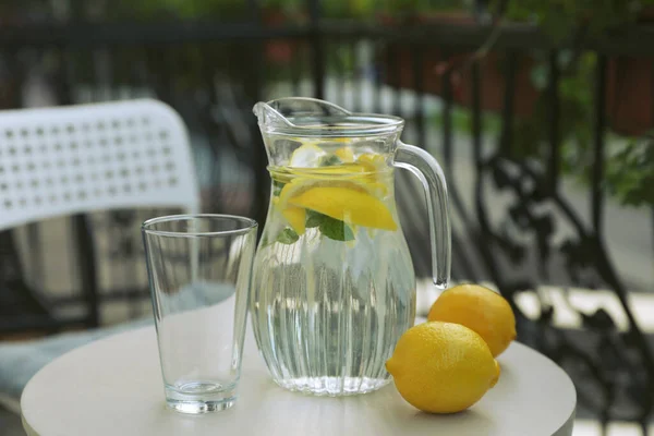 Jug with refreshing lemon water, glass and citrus fruits on light table outdoors