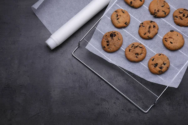 Roll of parchment baking paper and cooling rack with tasty cookies on dark grey table, flat lay. Space for text