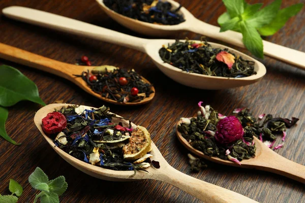 Spoons with dried herbal tea leaves and fruits on wooden table, closeup