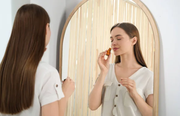 Young woman smelling essential oil near mirror indoors
