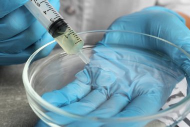 Doktor, gri masada şırıngadan petri kabına idrar damlatıyor.