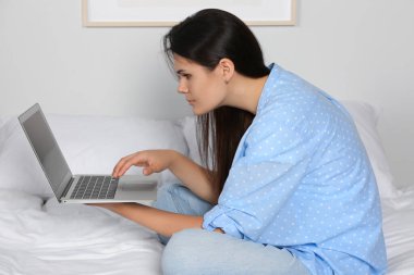 Evde yatağında otururken dizüstü bilgisayar kullanan kötü duruşu olan genç bir kadın. Skolyoz belirtileri