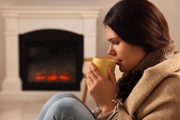 Young woman with cup of tea relaxing near fireplace at home. Space for text