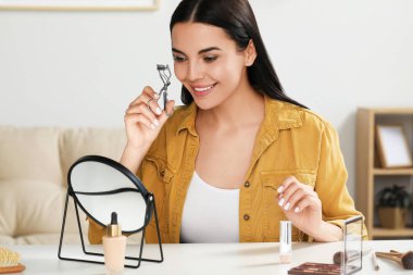 Güzel genç bir kadın, evde makyaj yaparken kirpik bigudisi kullanıyor.