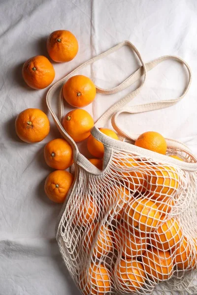 Net bag with many fresh ripe tangerines on white cloth, flat lay