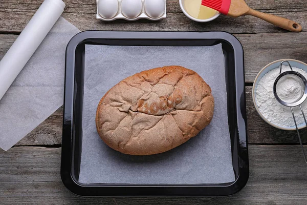 Parchment paper, baking pan with tasty homemade bread and different ingredients on wooden table, flat lay