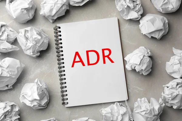 Alternative dispute resolution. Notebook with abbreviation ADR surrounded by paper balls on light grey table, flat lay