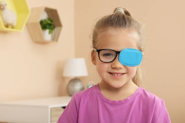 Girl with eye patch on glasses in room, space for text. Strabismus treatment clipart
