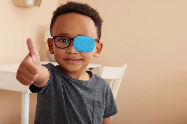 African American boy with eye patch on glasses showing thumb up in room, space for text. Strabismus treatment