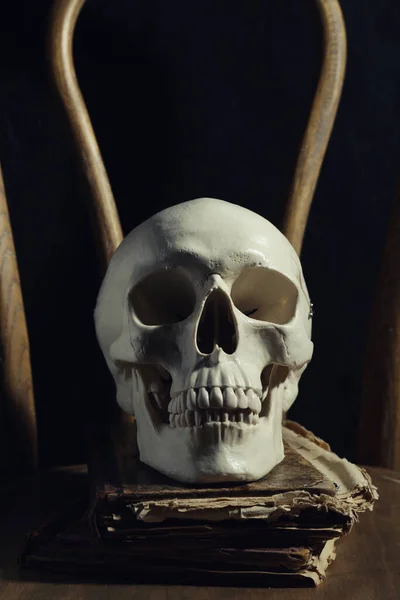 Human skull and old books on wooden chair against black background