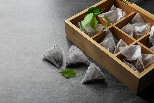 stock image Wooden box with paper tea bags and leaves on dark grey table, space for text