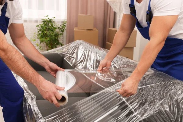 Workers wrapping sofa in stretch film indoors, closeup