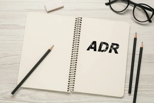 Alternative dispute resolution. Notebook with abbreviation ADR, pencils, eraser and glasses on white wooden table, flat lay