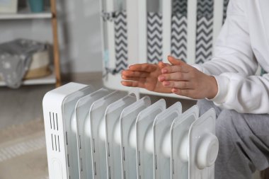 Genç bir kadın modern elektrikli ısıtıcının yanında ellerini ısıtıyor.