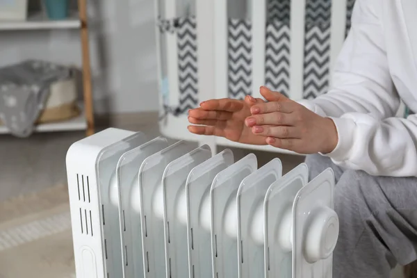 Young woman warming hands near modern electric heater indoors, closeup