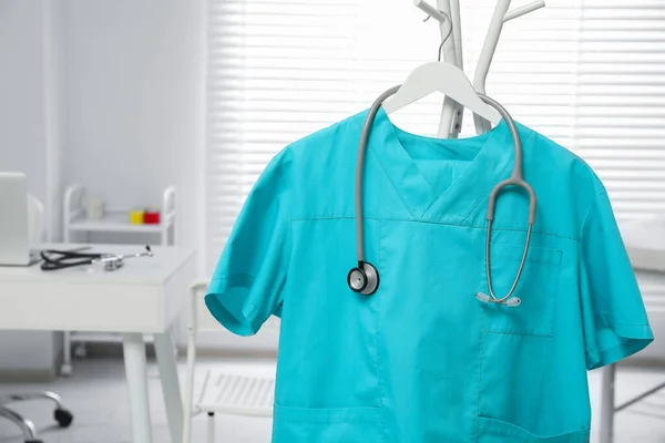 Turquoise medical uniform and stethoscope on rack in clinic, closeup. Space for text