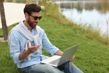 Göl kenarında yeşil çimlerde dizüstü bilgisayarı olan güneş gözlüklü bir adam.
