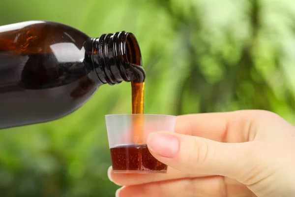 Woman pouring syrup from bottle into measuring cup on blurred background, closeup. Cold medicine