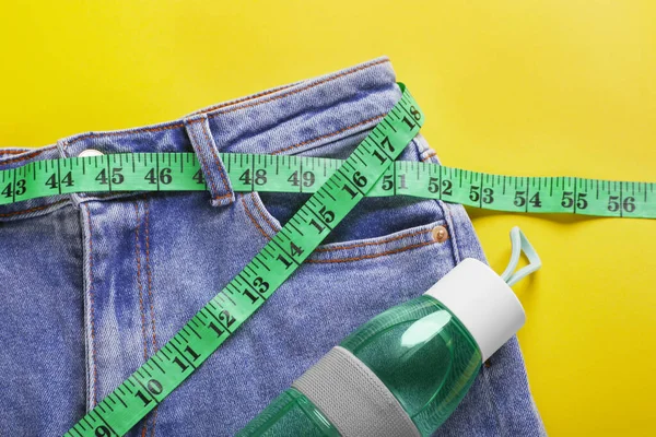 Jeans with measuring tape and bottle of water on yellow background, flat lay. Weight loss concept