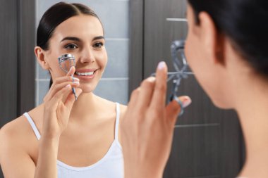 Güzel genç bayan banyoda aynanın yanında kirpik bigudisi kullanıyor.