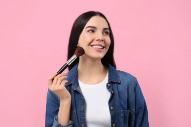 Güzel bir kadın pembe arka planda fırçayla makyaj yapıyor.