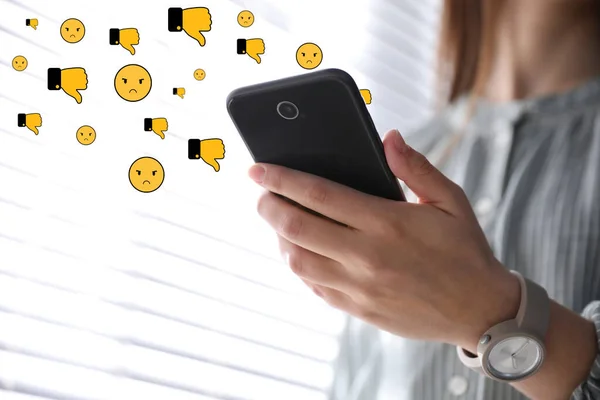 Social media dislike reaction. Woman using mobile phone indoors, closeup. Thumbs down and angry face emoji illustrations over gadget