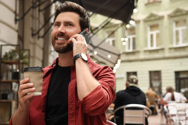 Handsome man with cup of coffee talking on smartphone outdoors, space for text