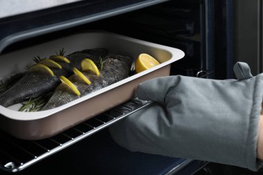 Kadın deniz levreği, limon ve biberiye ile pişirme tepsisini fırına koyuyor, yakın plan.