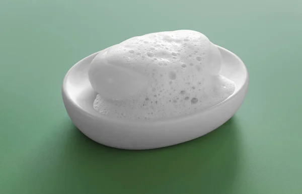 Dish with soap bar and fluffy foam on green background