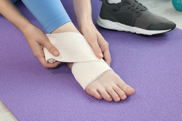 Woman wrapping foot in medical bandage on yoga mat indoors, closeup