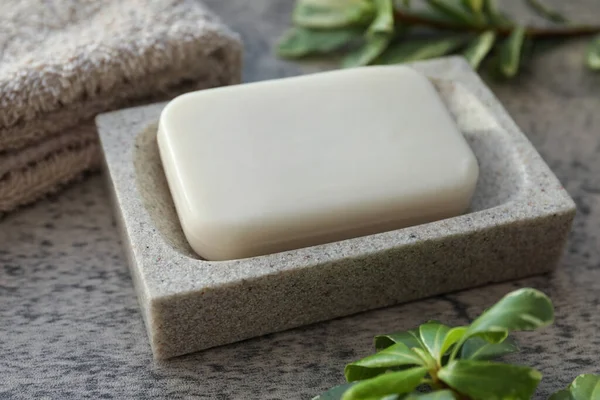 Dish with soap bar, terry towel and green plants on light grey textured table, closeup