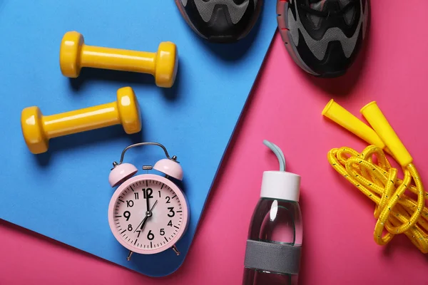 Alarm clock, bottle of water and sports equipment on pink background, flat lay. Morning exercise