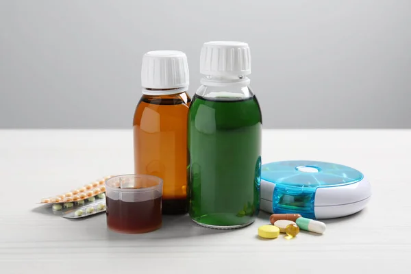 Bottles of syrup, measuring cup and pills on white table against light grey background. Cold medicine