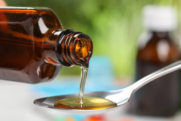Pouring syrup from bottle into spoon on blurred background, closeup. Cold medicine
