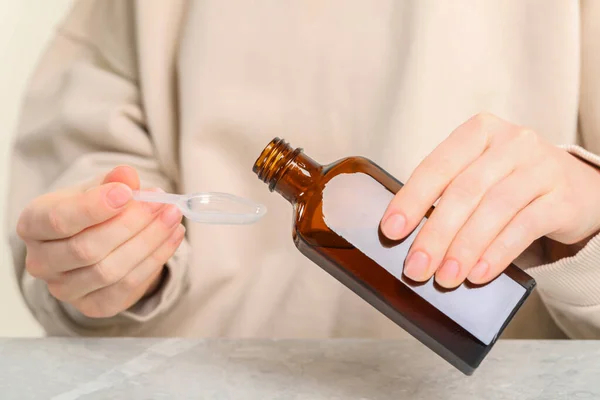 Woman pouring syrup from bottle into dosing spoon at table, closeup. Cold medicine
