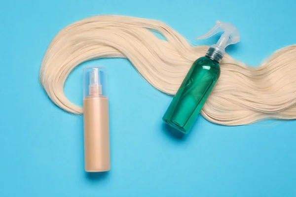Spray bottles with thermal protection and lock of blonde hair on light blue background, flat lay