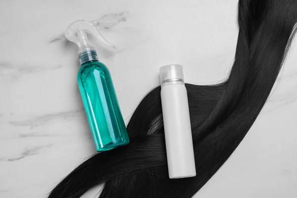 Spray bottles with thermal protection and lock of brunette hair on white marble table, flat lay