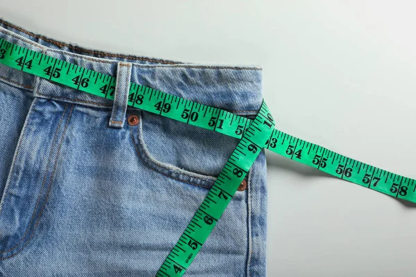 Jeans with measuring tape on light grey background, top view. Weight loss concept